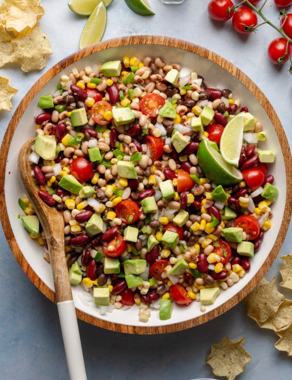 Mixed cowboy caviar in a large bowl with a wooden spoon, showing vibrant beans, corn, tomatoes, jalapeños, and diced avocado.