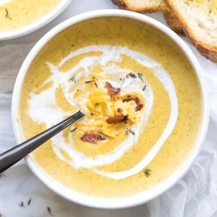 a white bowl with yellow soup and white cream swirl topped with shredded cheese and bacon crumbles